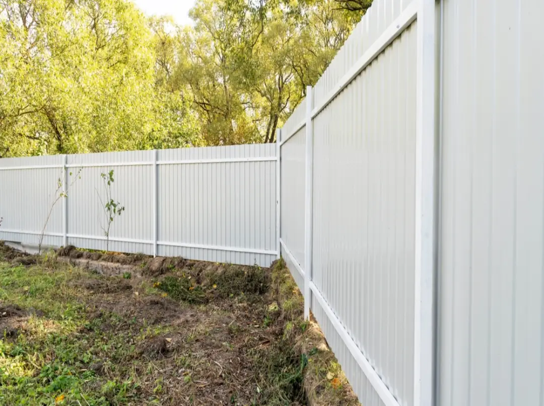 Clean white vinyl privacy fence and gates installed along a residential property perimeter with lush trees in the background.
