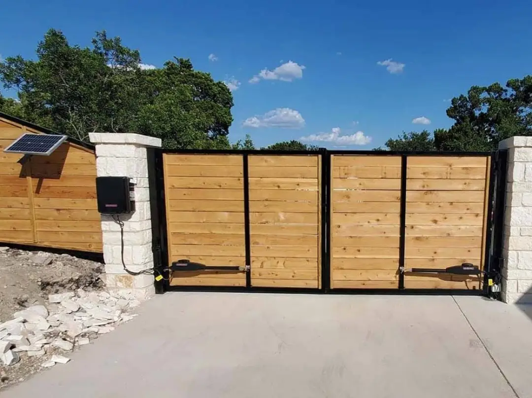 Automated dual-swing driveway fence and gates with horizontal wood slats and black metal frame on concrete driveway.